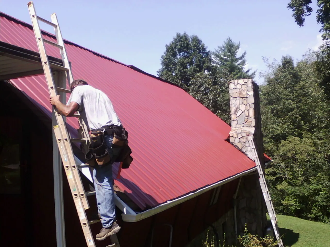 Roofer on a ladder beside a red metal roof and stone chimney — Soffit & Fascia Repair by Roofing Brothers in Leon
